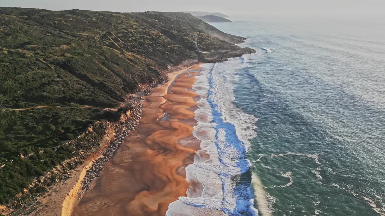 Clip aerea al rallentatore di Praia do Salgado a Nazaré che cattura le onde che si infrangono contro la scogliera frastagliata e la spiaggia sabbiosa durante una giornata luminosa. Perfetto per progetti di viaggio e natura