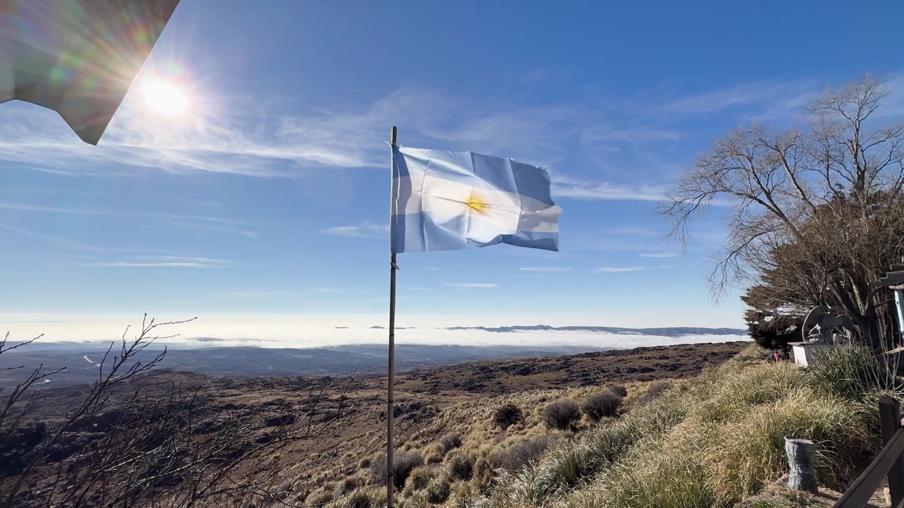 Argentinsk flagga vajar ovanför Andernas fot nära Quebrada de Zonda, San Juan, Argentina, med utsikt över Anderna, torra högland och ett vidsträckt bergslandskap, statisk bild.