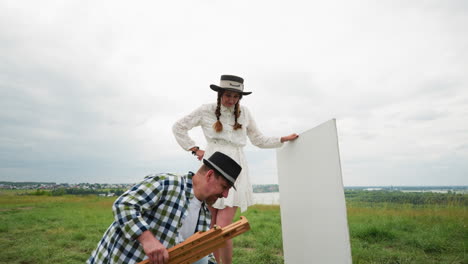 artistic couple in a field setting up canvas and easel, man in plaid shirt and hat, woman in white dress. the sky is casting a gentle, diffused light on the scene, which adds to the tranquil mood