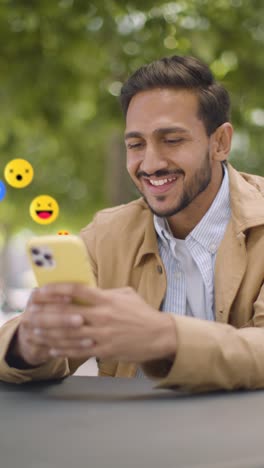 video vertical de un hombre sonriente sentado en una mesa al aire libre mirando un teléfono móvil con emojis de gráficos en movimiento que muestran múltiples notificaciones de redes sociales gustando y reaccionando al contenido en línea 1