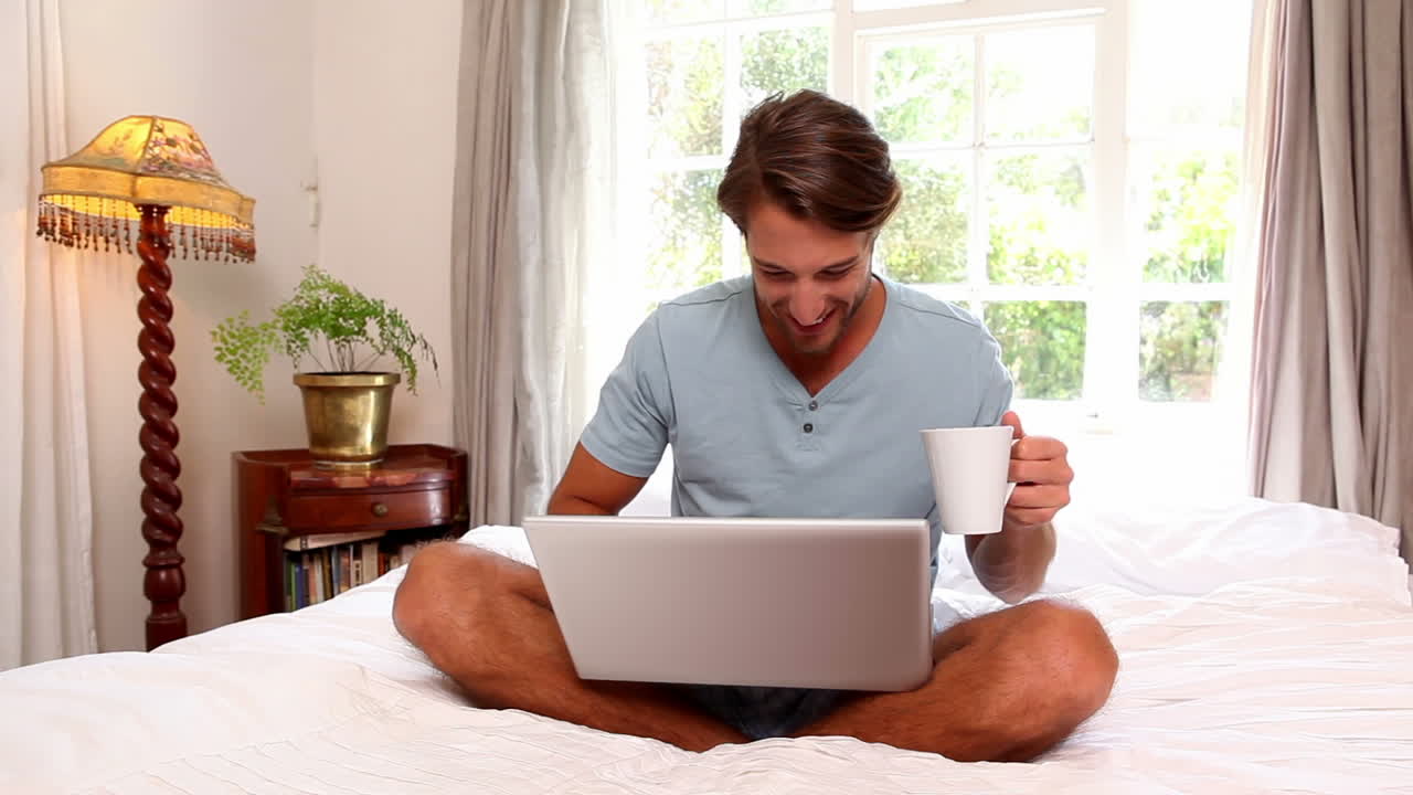 hombre guapo sentado en la cama usando una taza con una computadora portátil