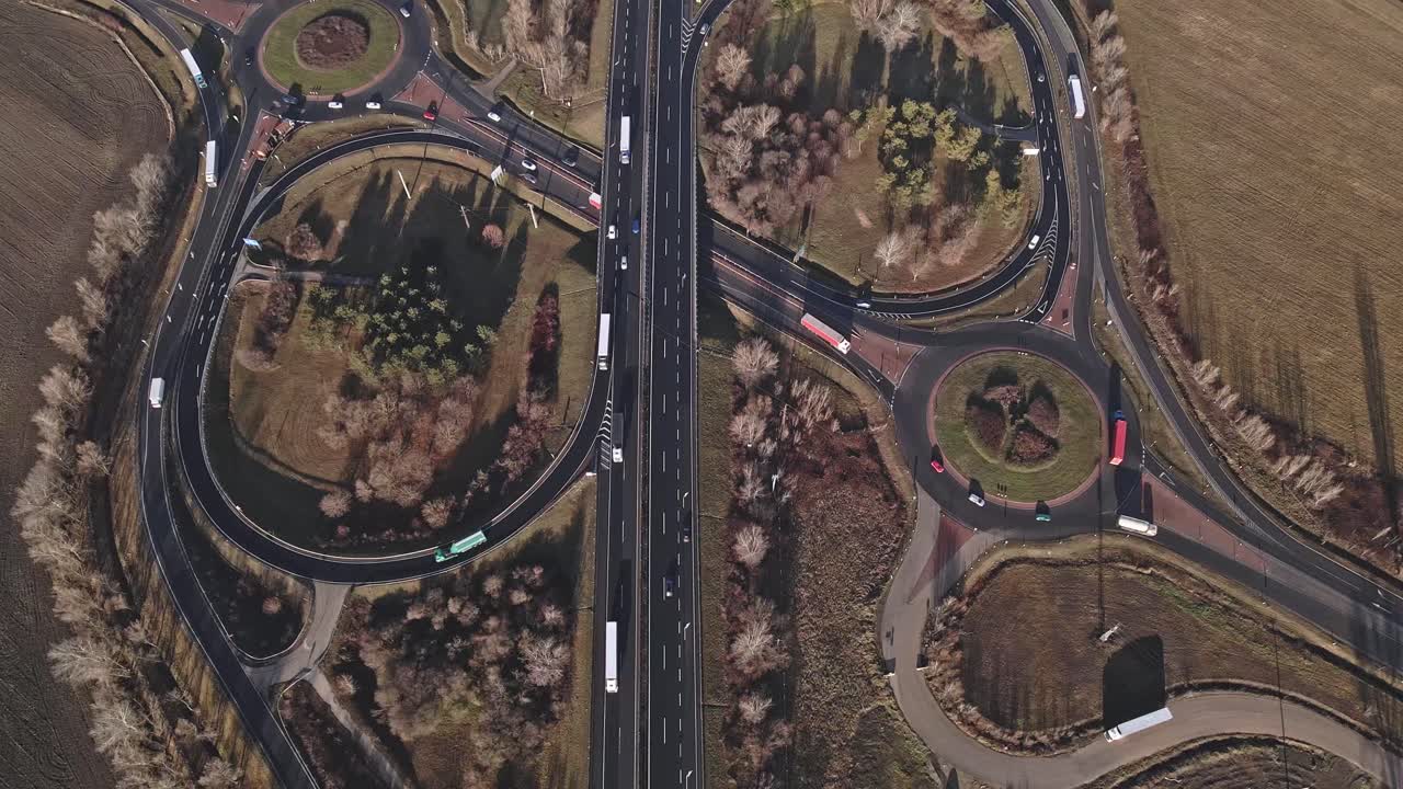 A drone-captured video of a highway with two large roundabouts surrounded by green spaces and trees. Flying over the moving cars and trucks on the highway.