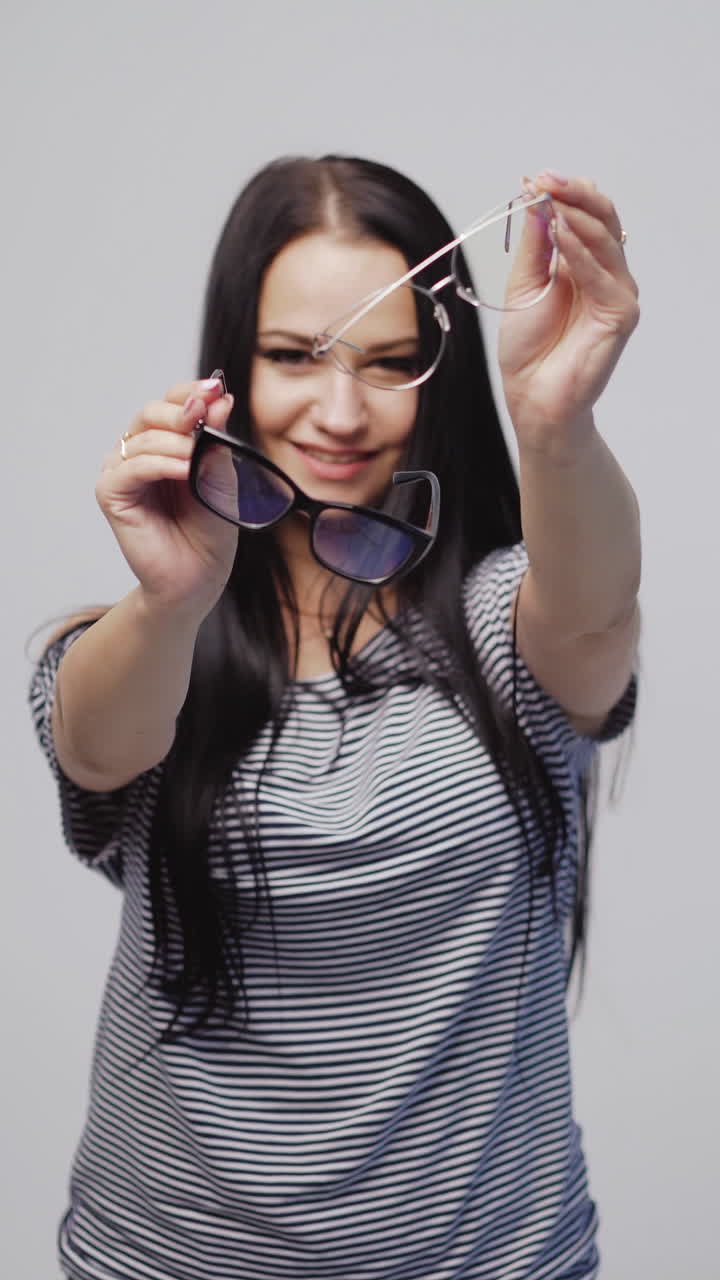 beautiful woman in a striped dress is showing two models of eyeglasses with a black and white rims on a gray background. Vertical video