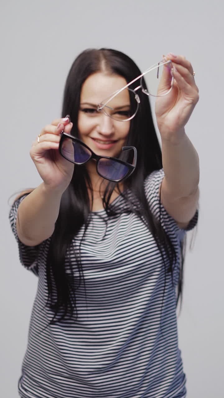 beautiful woman in a striped dress is showing two models of eyeglasses with a black and white rims on a gray background. Vertical video