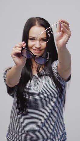beautiful woman in a striped dress is showing two models of eyeglasses with a black and white rims on a gray background. vertical video