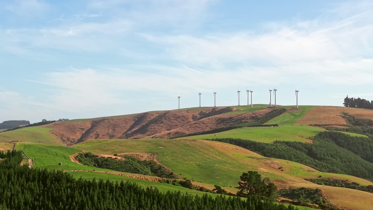 Smooth drone flight approaching and rising toward tall white turbines on a green hillside, revealing scenic countryside views of New Zealand’s rural landscape