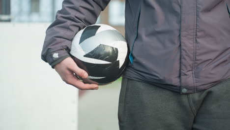 close-up of someone walking into a sports environment with hand holding black and white soccer ball with focus on player s hand, showing athletic wear and part of body, urban background