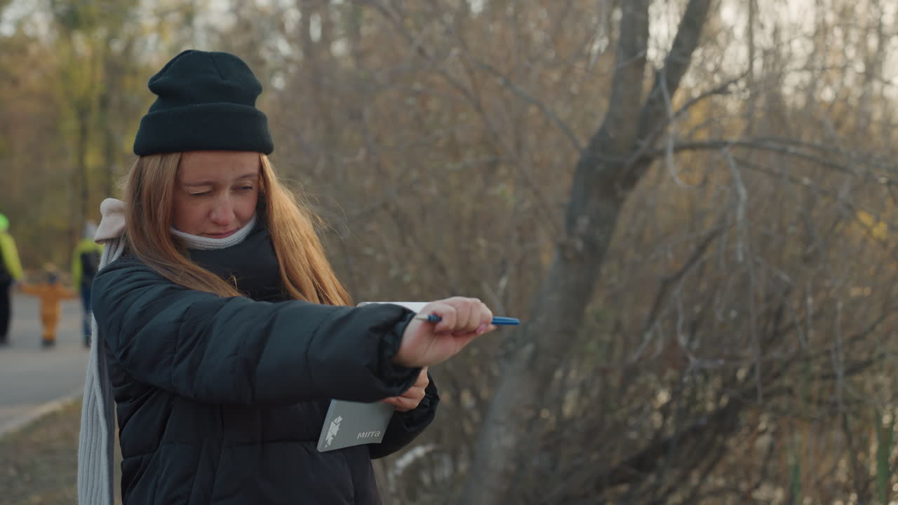 Donna bianca che punta la penna mentre registra all'aperto, berretto e mascherina sotto il mento, taccuino tenuto fermo vicino al sentiero del fiume, luce autunnale vivace e rami spogli, energia da reporter determinata e stile documentaristico.