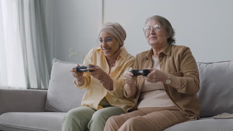 feliz mujer árabe de mediana edad y anciana jugando videojuegos juntos y divirtiéndose mientras se sientan en el sofá en casa