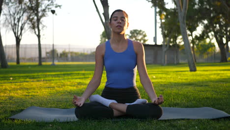 An-attractive-young-hispanic-woman-on-a-yoga-mat-sitting-in-a-lotus-meditation-pose-in-the-park-at-sunset