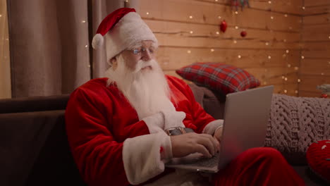 Porträt-Des-Weihnachtsmanns-Mit-Bart-Und-Brille,-Der-Am-Heiligabend-Mit-Einem-Laptop-Sitzt-Und-Geschenke-Im-Online-Shop-Bestellt