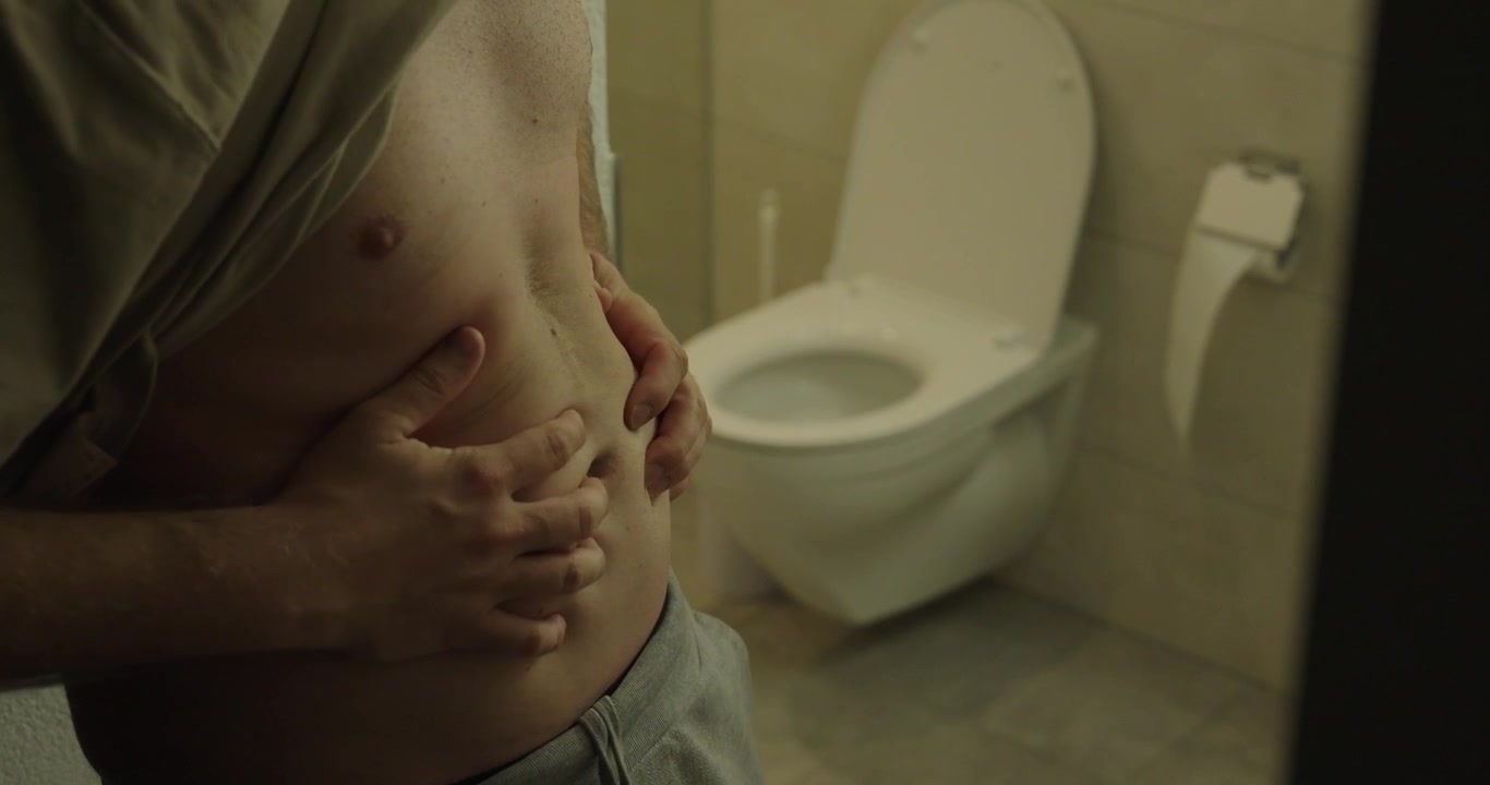 Person holding their stomach while standing near a toilet, illustrating abdominal pain, digestive discomfort, constipation, or gastrointestinal health issues in a home bathroom setting.