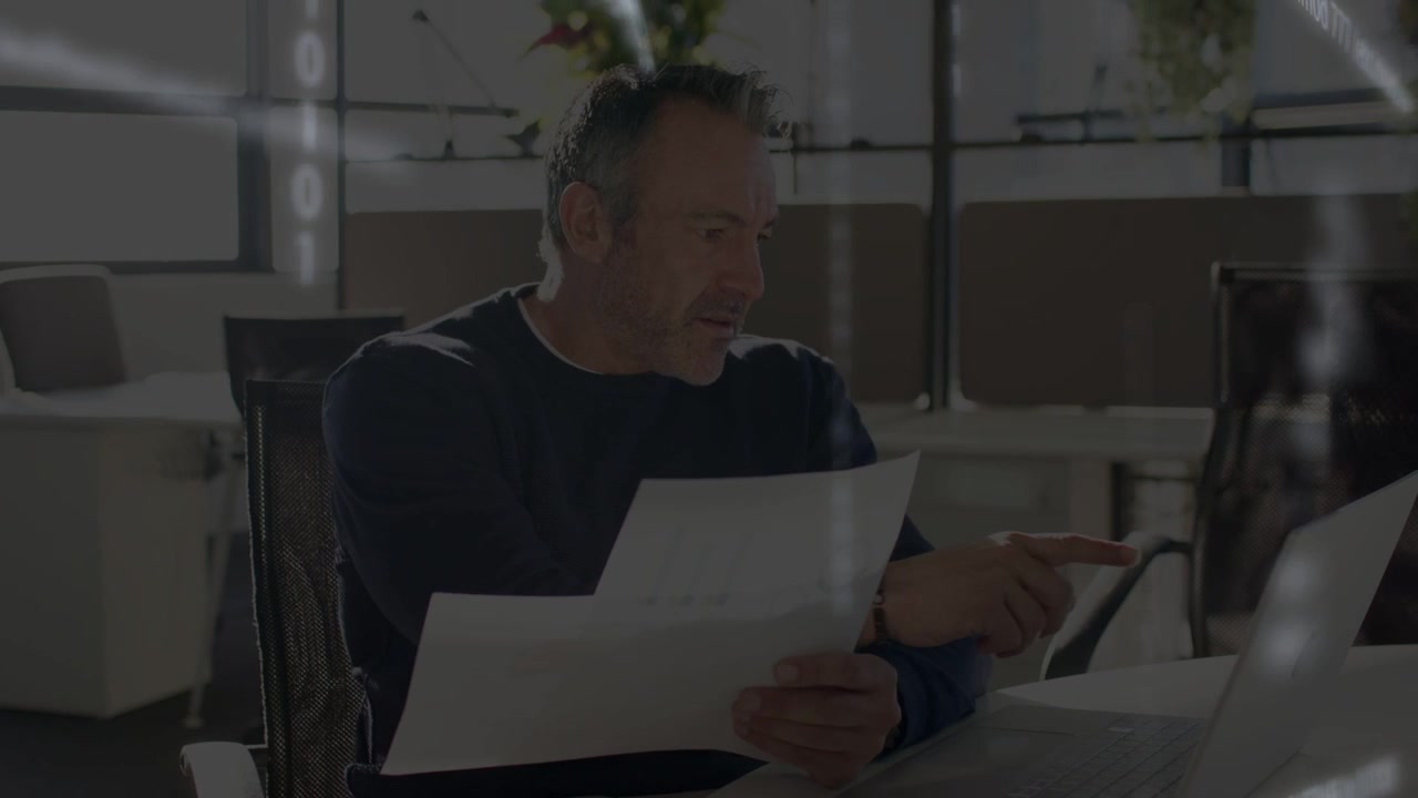 Homem lendo papéis e depois apontando para o laptop, iniciando sobreposições binárias enquanto analisa dados financeiros.