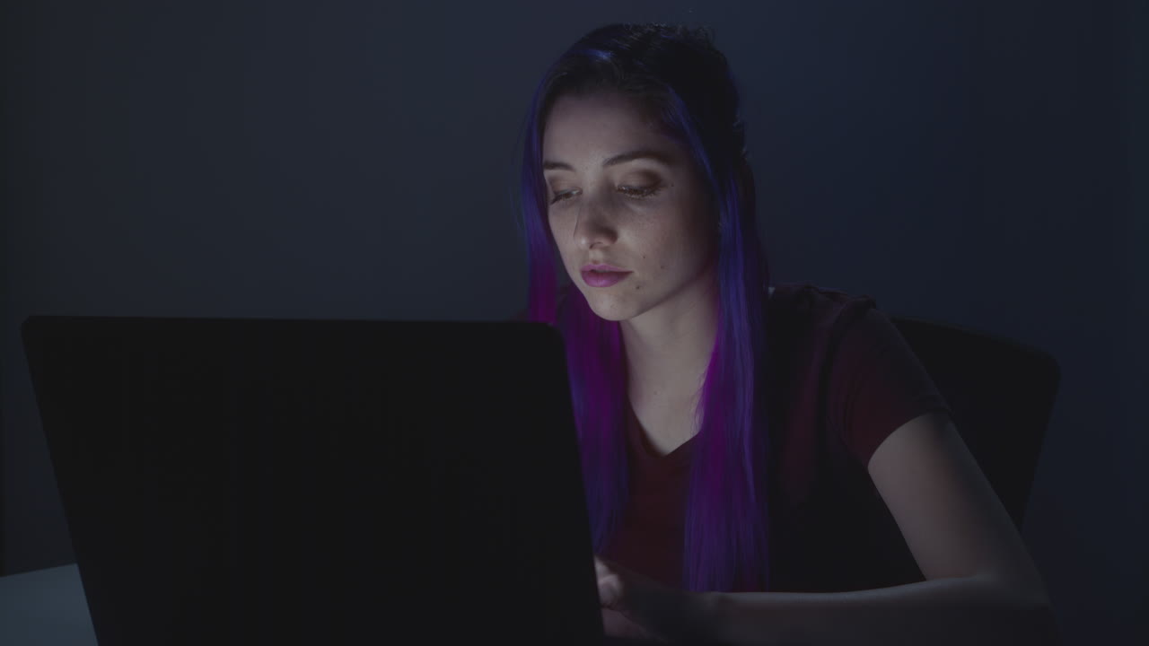 Young girl working in her laptop, the room is dark and the screen lights her face