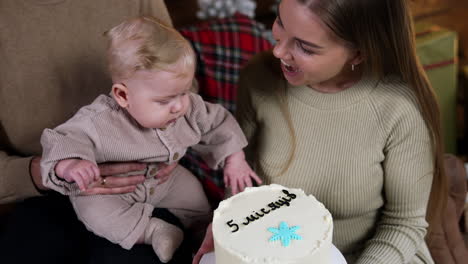 Cute little baby staring on the cake in his mom's hands. Family celebrates five months of child's life.
