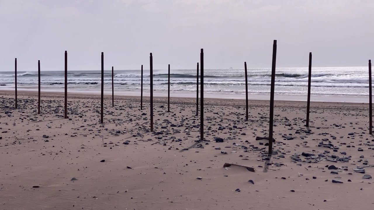 Postes de madeira alinham uma praia arenosa e de seixos, com ondas quebrando na costa e falésias surgindo ao longe.