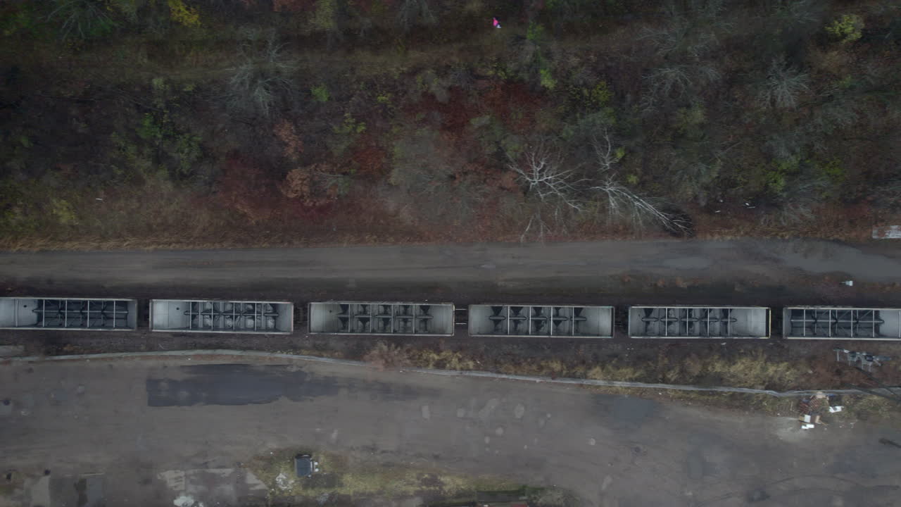 Aerial drone tracking shot with a long freight train passing through an industrial area bordered by trees and scattered debris