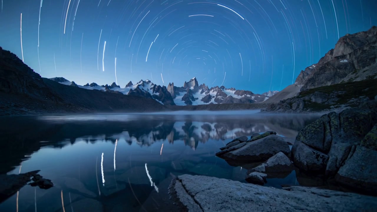 霧がかかった花崗岩の岩々がある湖に、山頂の上を回転する星々が映り込むタイムラプス映像。