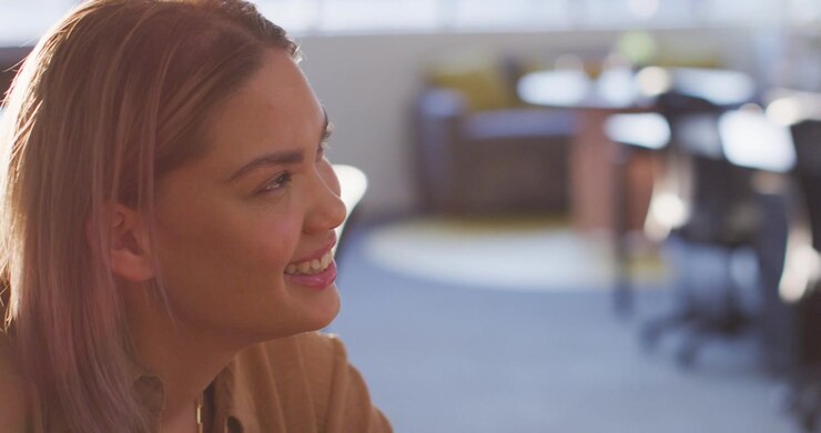 Woman sitting at table in open office, listening, smiling and laughing at coworker joke, copy space