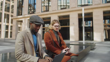 two multiethnic male colleagues speaking and using gadgets outdoors