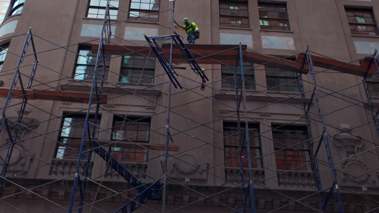 A low angle view, looking up at workers erecting scaffolding onto a building façade in New York City on a cloudy day. The camera is stationary tilted upwards.