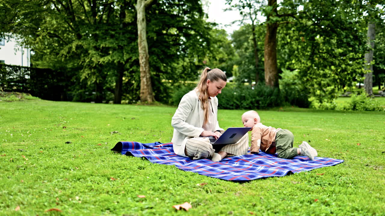 En ung mor sidder på et picnictæppe i en grøn park med sin lille søn. Den lille søn løber ved siden af sin mor og kigger interesseret på den bærbare computers skærm. Den unge mor arbejder fjernstyret.