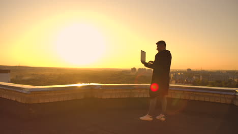 a male stockbroker freelancer stands on a rooftop at sunset with a laptop and types on a keyboard with his fingers looking at the cityscape from a bird's eye view.