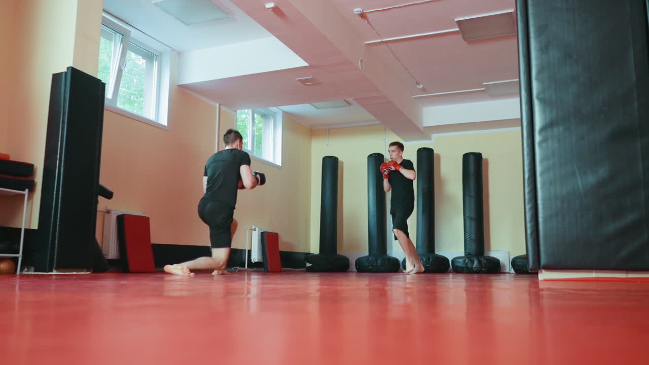 Two boxers sparring in martial arts gym practicing stance with gloves on red mat floor surrounded by punching bags under daylight from windows during combat training session focusing on skill technique