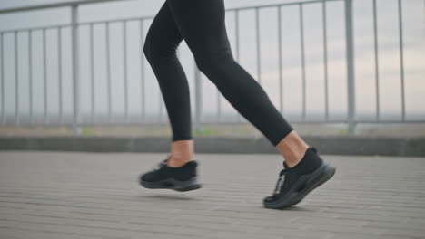 close-up view of woman s legs jogging for morning fitness in black leggings and black sneakers, showing active workout routine on paved path with iron fence in background