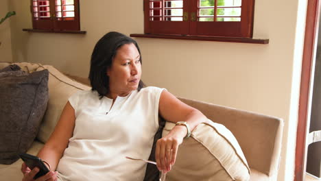 Woman relaxing on sofa at home, browsing smartphone with focused expression