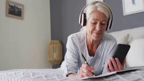 Happy-senior-caucasian-woman-laying-on-bed-in-bedroom,-using-smartphone-and-wearing-headphones