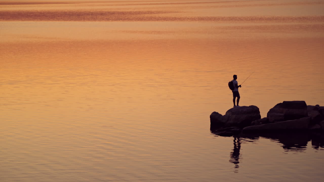 Silhouette of a fisherman with rod on the orange water background. Man with fishing rod is standing on a rock at a beautiful sunset in water.