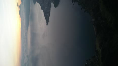 Drone-portrait-view-in-Guatemala-flying-over-a-blue-lake-surrounded-by-green-mountains-at-sunrise-on-a-cloudy-day-in-Atitlan