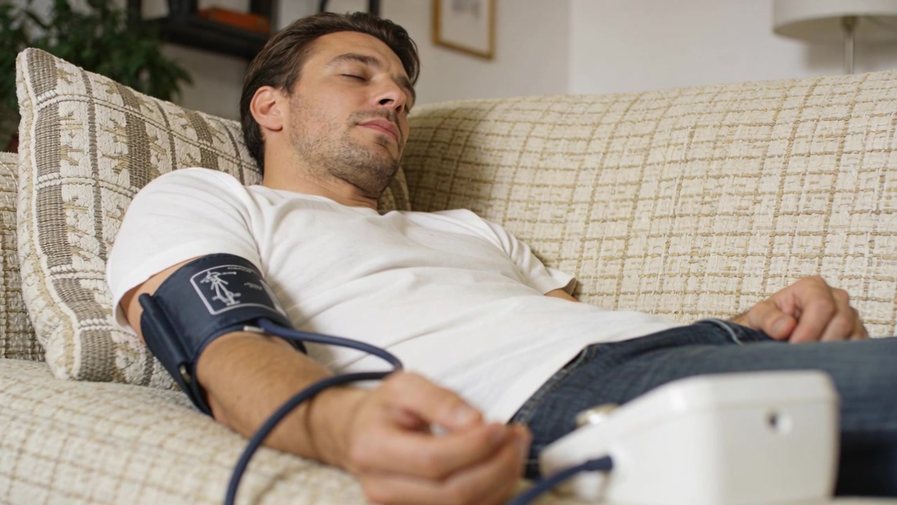 Uomo sdraiato in maglietta bianca e jeans che controlla la pressione sanguigna a casa, con il bracciale che si gonfia per monitorare.