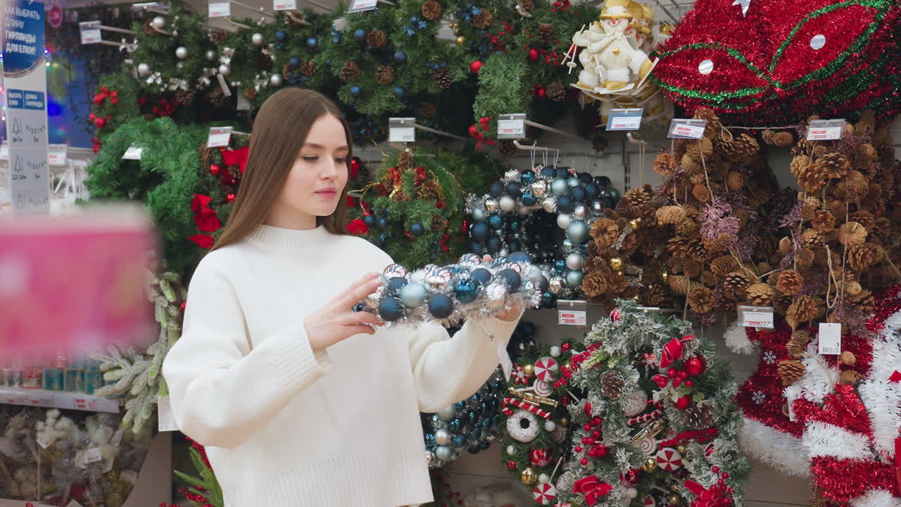 Young woman in white sweater picks up festive Christmas decor from a display, then playfully places it on her head, she enjoys shopping in a well-decorated holiday store, surrounded by ornaments