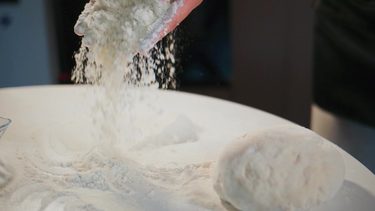 Hands knead white dough on floured table. Flour dusts air as hands work. Scene shows baking preparation. Ideal for recipes, food blogs, or cooking tutorials. Focus on tactile, hands-on baking process