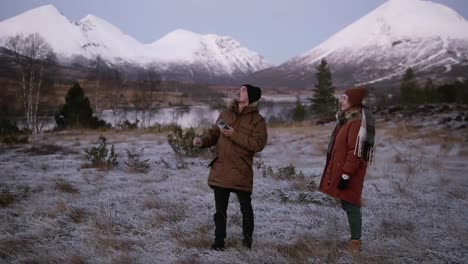 una pareja caucásica lanza drones en el campo frente a las montañas con picos nevados al fondo. hombre operando el helicóptero, mujer parada cerca. campo nevado alrededor