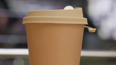 close up brown takeaway coffee cup with open lid placed on table with blurred bokeh background featuring warm lighting and faint moving reflections behind glass in cozy indoor setting
