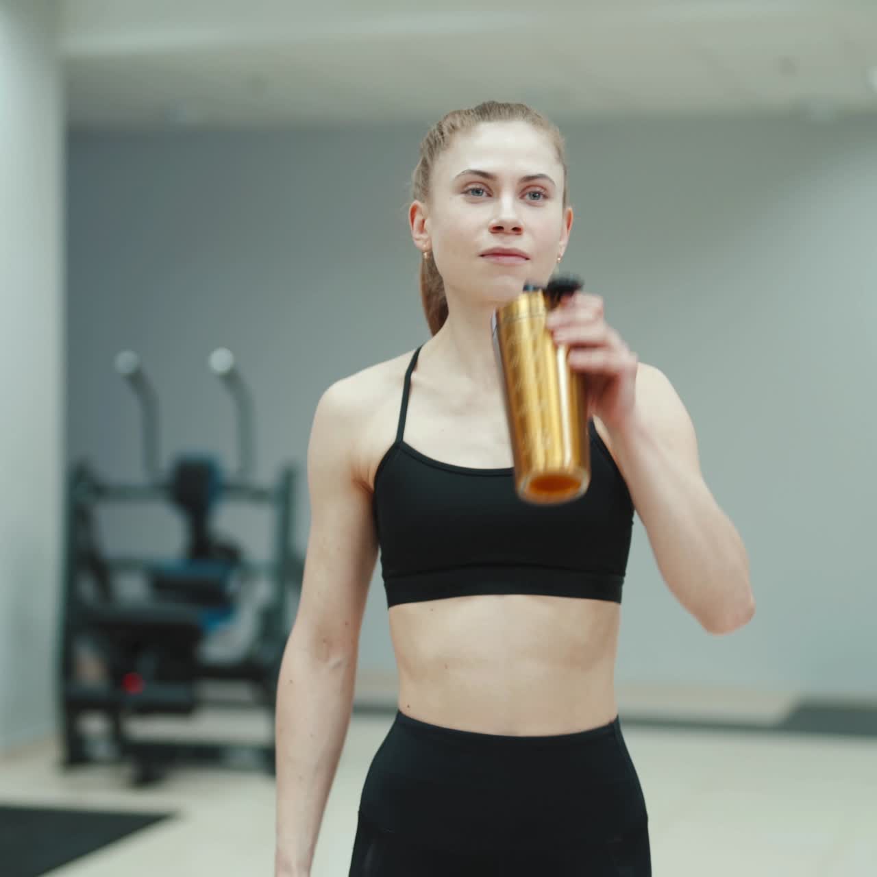 A slender woman in a black sports top and with her hair stuck in the tail is drinking water from a shaker and walking through the gym. Blurred Background