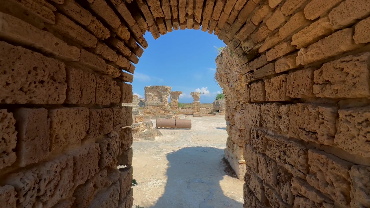 Arco in pietra che incornicia un cortile aperto tra le rovine di Cartagine, con una forte luce di mezzogiorno e ombre profonde. Tunisia.