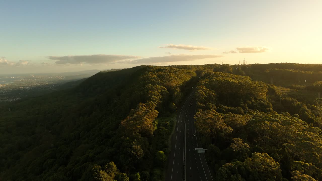 Rugged ridges of the Illawarra Escarpment lit by golden hour light near the coast, aerial tracking follows highway on ridge