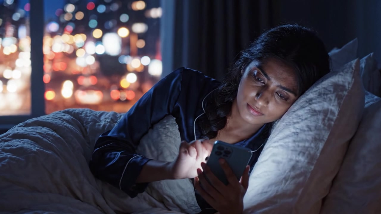 Una giovane donna assorta nel suo dispositivo mobile mentre si rilassa a letto, a tarda notte, illuminata da luce soffusa e con vista sulla città notturna.