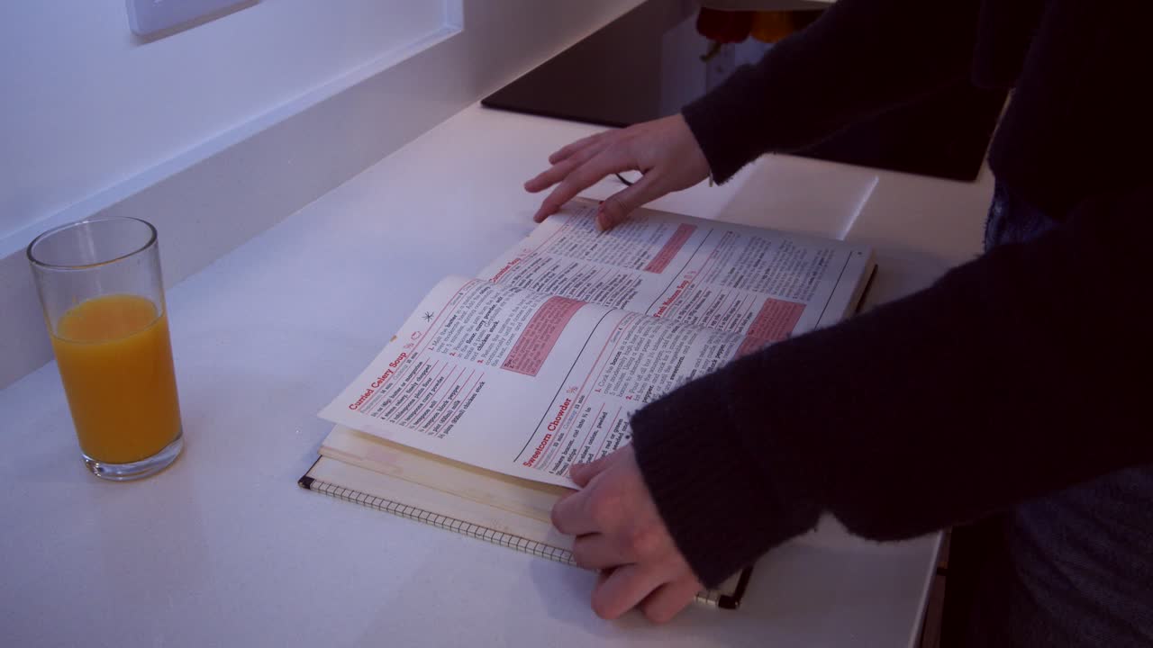 modelo femenino mirando a través de un libro de cocina en una encimera de cocina, luego cerrando el libro y caminando