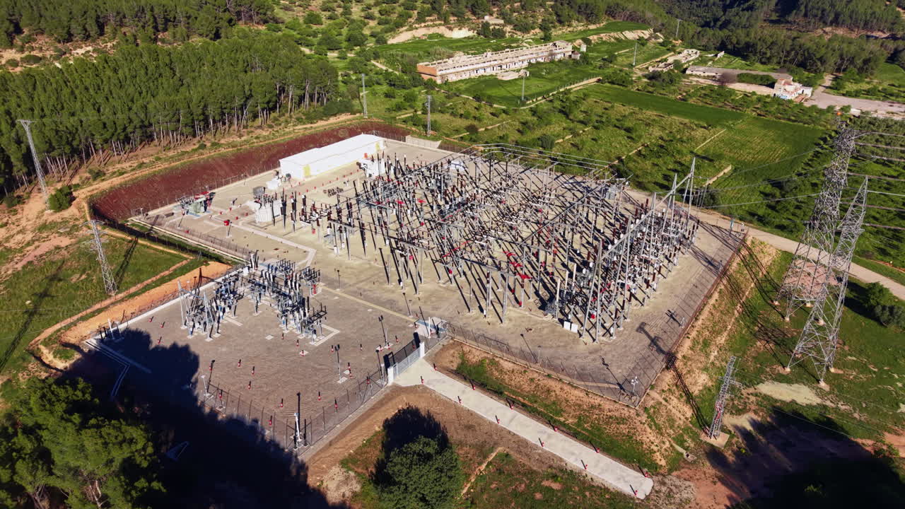 Overhead view of a large electrical substation surrounded by green forest and farmland, featuring transformers, transmission towers, and complex wiring infrastructure