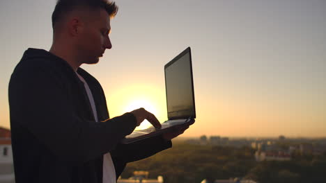 Hacker-Usando-Una-Computadora-Portátil-En-La-Azotea-Con-Vista-A-La-Ciudad-Y-Gráfico-De-Divisas.-Concepto-De-Piratería-Y-Estadísticas.-Un-Hombre-Al-Atardecer-En-Cámara-Lenta-Escribiendo-Código-De-Software-En-Una-Computadora-Portátil