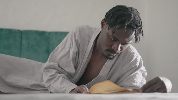 Man in white robe lying on bed, deeply focused on reading large book, turning pages slowly. Soft natural light filters through window, creating peaceful and reflective atmosphere in indoor space
