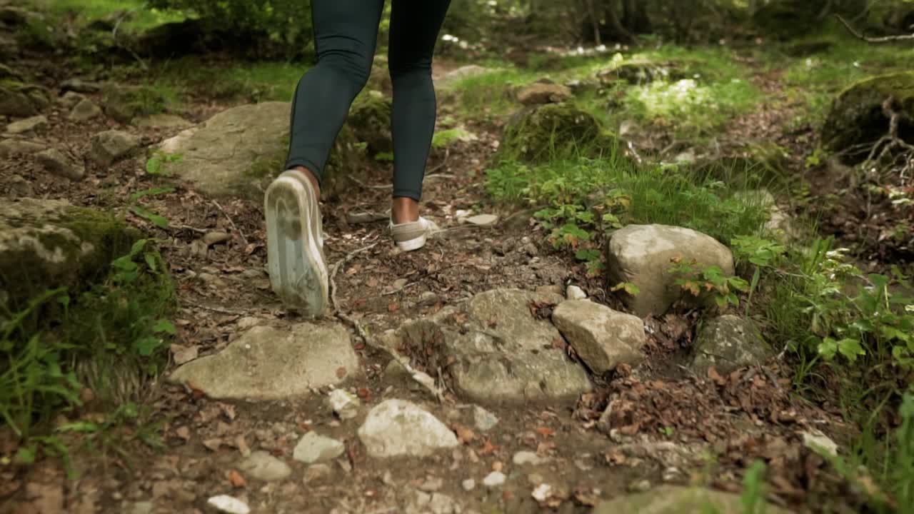 mujer en forma con muslos negros usando zapatillas ligeras caminando por un camino de tierra rocosa a través de los árboles del bosque en una montaña
