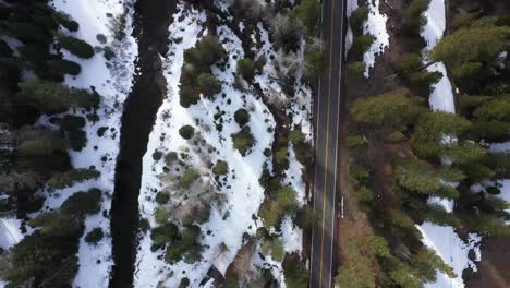 aerial view from drone looking straight down at winter stream, tall trees and a 2 lane road going through the mountains following a light snowfall