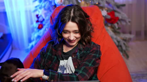 joyful person relaxes with cat. a joyful individual sits on a bean bag, using a laptop while a cat curiously approaches in a cozy, decorated room