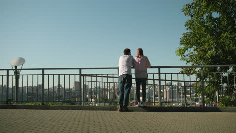 back view of two friends leaning on iron railing overlooking vibrant urban cityscape while engaging in conversation, lady stands on pavement while boy gestures, surrounded by greenery